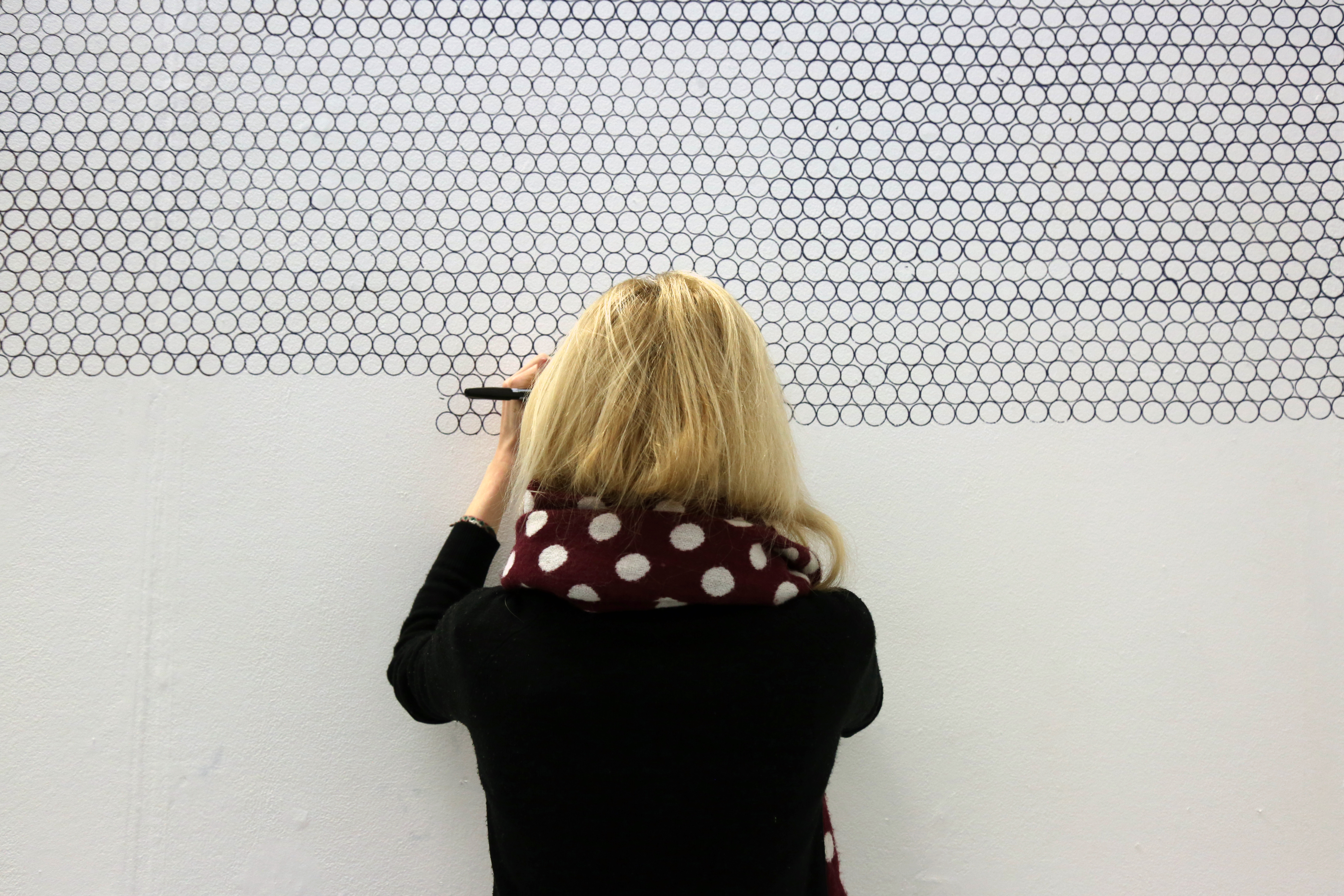 Amelia repetitively draws round a penny coin in continuous lines with a black pen onto a white wall. She is stood with her back to the camera wearing a black dress and is also wearing a scarf that mimics the circles she is creating.