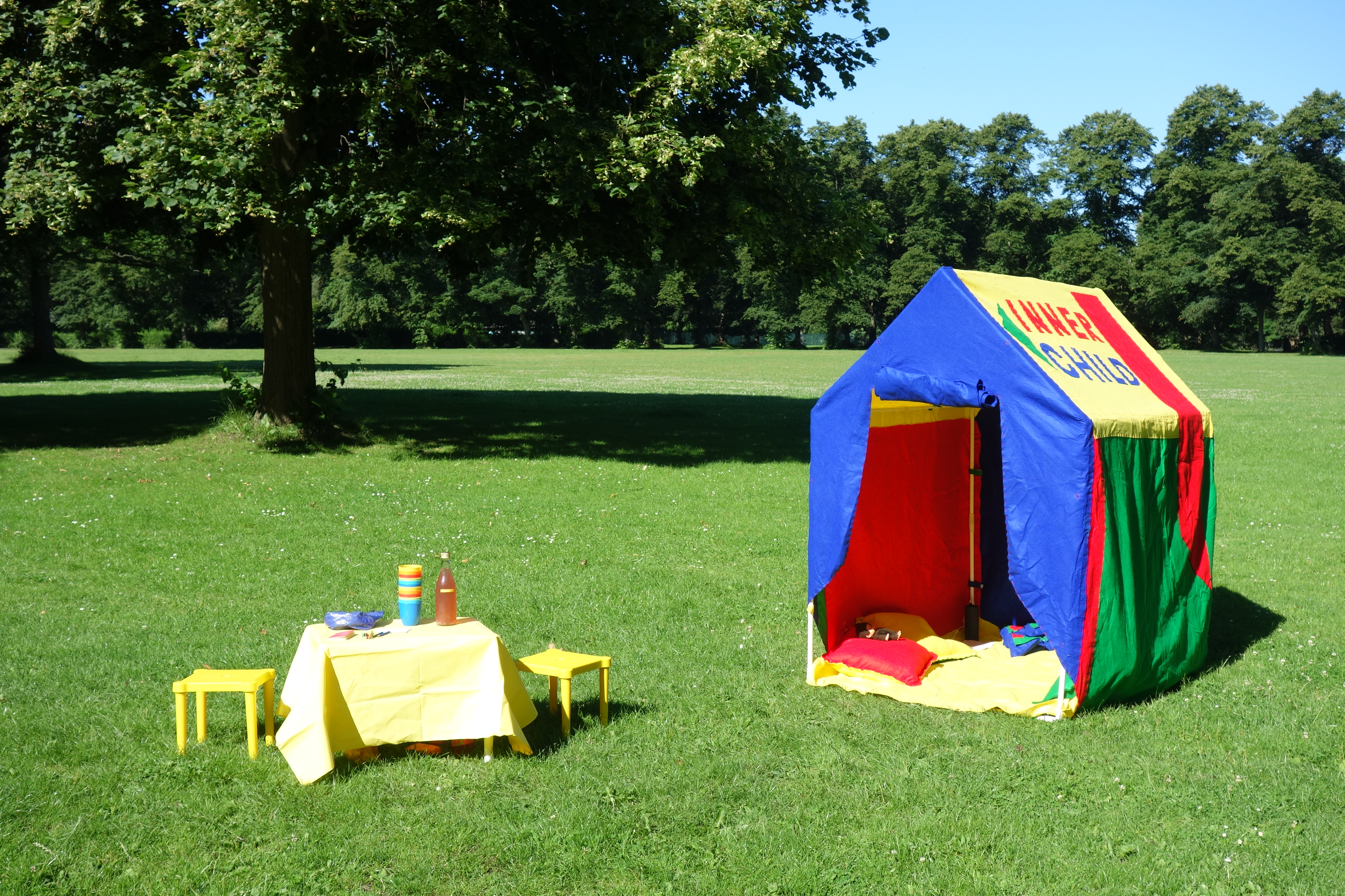 In a Park, on a sunny day, there is a felt hand-made children hut in green, red, yellow and blue, next to it a children plastic table and two chairs, in yellow.