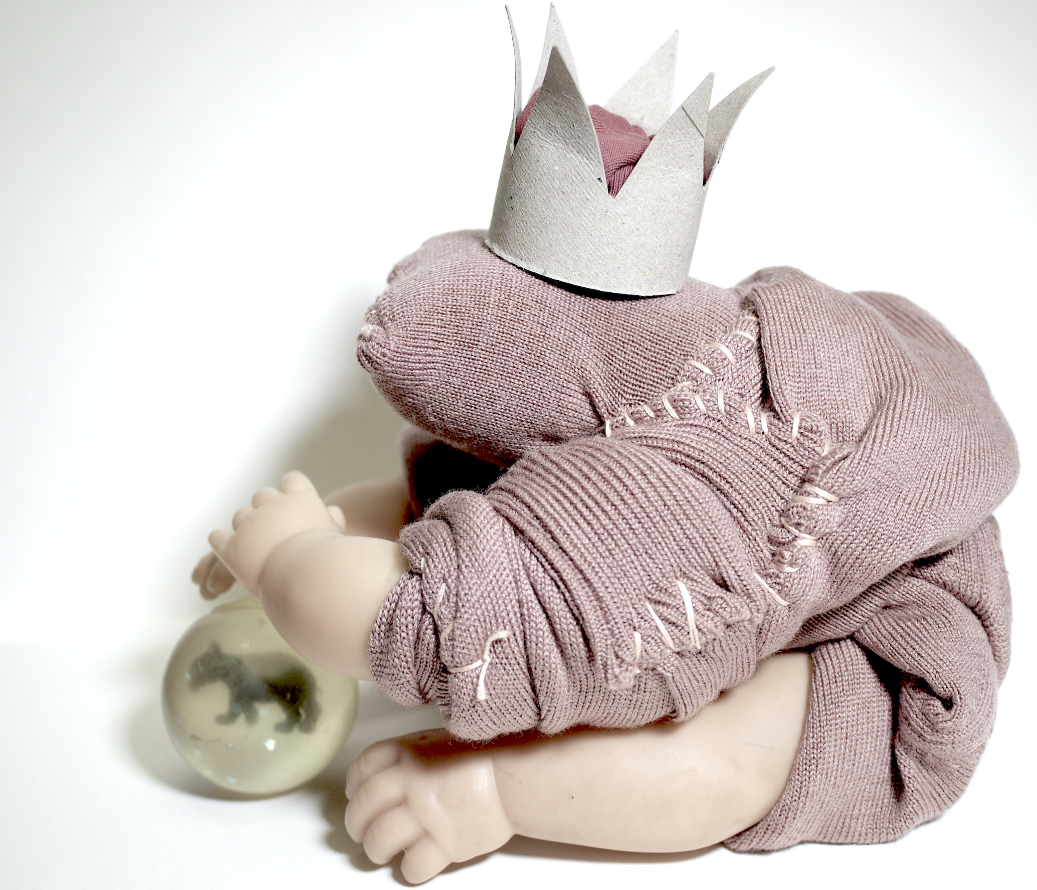 The sculptural work was created during the first Covid-19 lockdown, whereas the artist could only use existing materials from her home studio. An old dusty pink jumper was used as the body sewn together and four doll hands were used as the legs. The frog is holding an old translucent bouncy ball with a small plastic animal embedded into it donated by the artist’s son. The crown the frog wears was created from a toilet roll as the documentation of the panic buying of toilet rolls at the time.